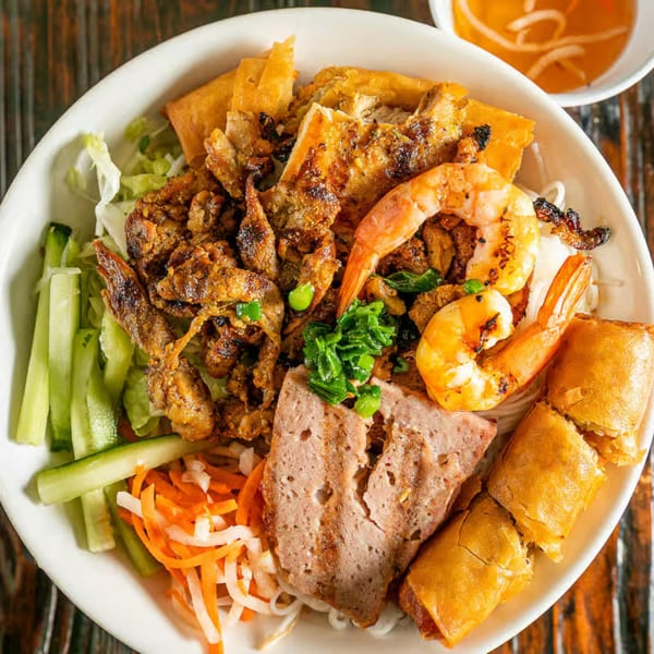 A large bowl of Vietnamese vermicelli noodles with grilled pork, shrimp, spring rolls, and vegetables.