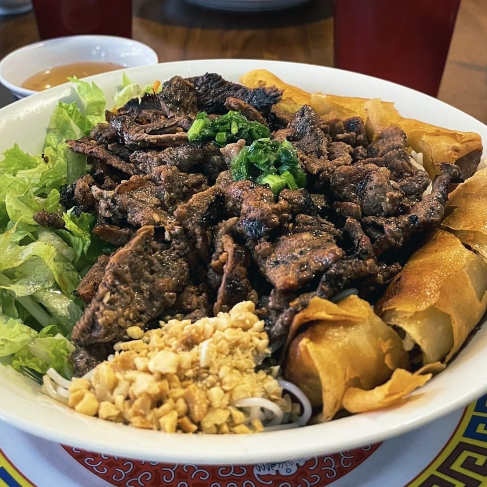 A large bowl of grilled beef, lettuce, peanuts, and crispy spring rolls, with a sauce bowl.